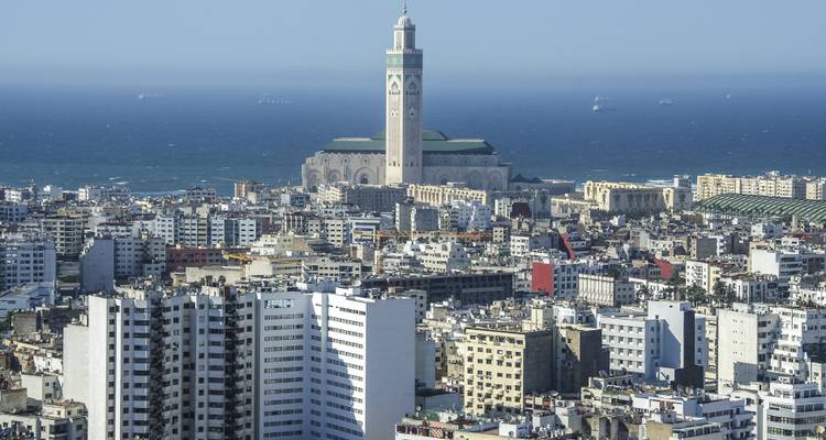 Amplio paisaje urbano de Casablanca con la Mezquita Hassan II elevándose sobre un denso conjunto de edificios modernos y el Océano Atlántico al fondo.