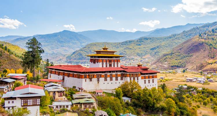 Forteresse-monastère de Rinpung Dzong avec des murs rouges et blancs située contre les contreforts himalayens boisés et un ciel bleu.