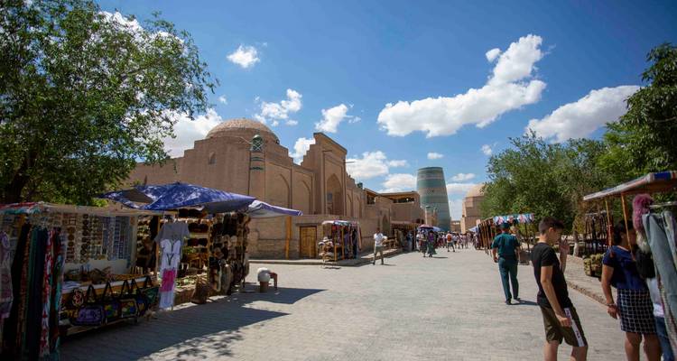 Rue piétonne animée à Khiva bordée d'étals d'artisanat, avec des coupoles ensoleillées et le minaret Kalta Minor au loin.