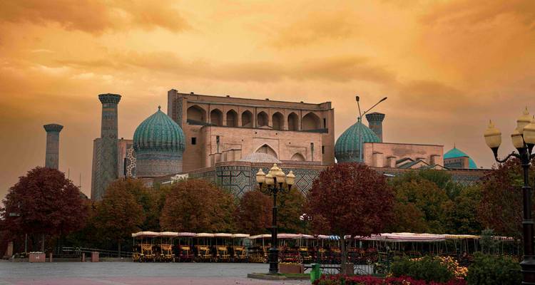 Place du Régistan à Samarcande sous un ciel dramatique doré du soir avec des arbres d'automne au premier plan.