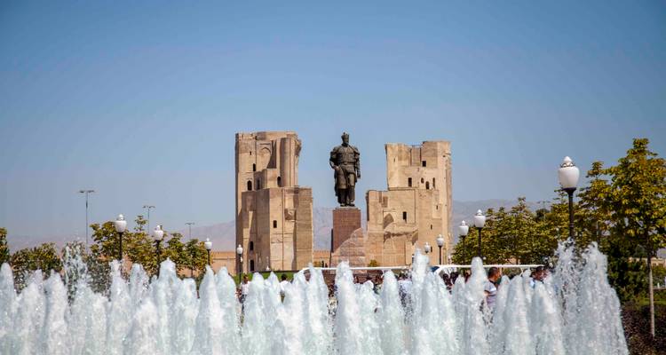 Des jets de fontaine encadrent une statue d'Amir Temur avec des murs de palais en ruines derrière sous un ciel bleu clair.