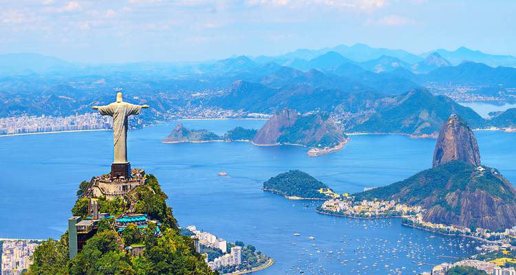 La icónica estatua del Cristo Redentor en la cima del Corcovado con vista a la Bahía de Guanabara y el Pan de Azúcar con un panorama costero expansivo.