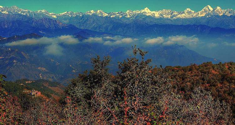 Una distante muralla nevada del Himalaya se alza sobre colinas ondulantes con nubes bajas flotando por debajo