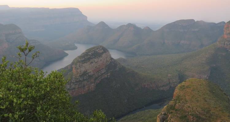 Vista brumosa del amanecer del Cañón del Río Blyde con escarpadas mesetas que se elevan sobre un río serpenteante y una meseta distante.