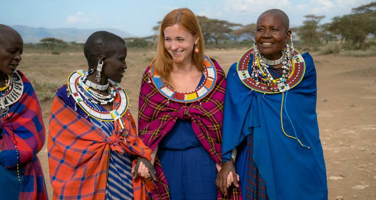 Glimlachende reiziger staat arm in arm met kleurrijk geklede Maasai vrouwen versierd met ingewikkeld kralenwerkop een open vlakte.