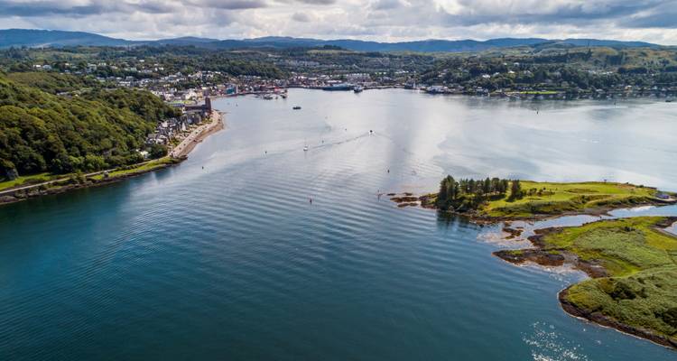 Luchtfoto van een Schotse kustplaats met een kalme baai, beboste heuvels en kabbelende bootsporen op blauw water.