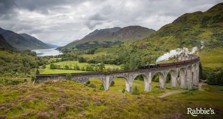 Dramatisch luchtfoto van een stoomtrein die het Glenfinnan Viaduct oversteekt te midden van weelderige Schotse heuvels, met een logo watermerk in de hoek.
