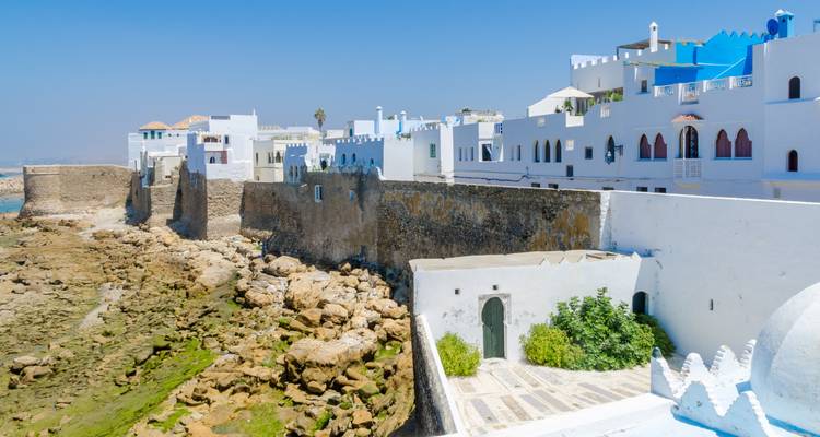 Maisons blanches et bleues en bord de mer bordant d'anciens remparts au-dessus d'un rivage rocheux.