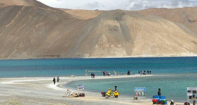Las aguas turquesas de un lago de gran altitud se encuentran con una lengua de arena donde pequeños grupos de turistas caminan y toman fotos; áridas colinas marrones se elevan detrás.