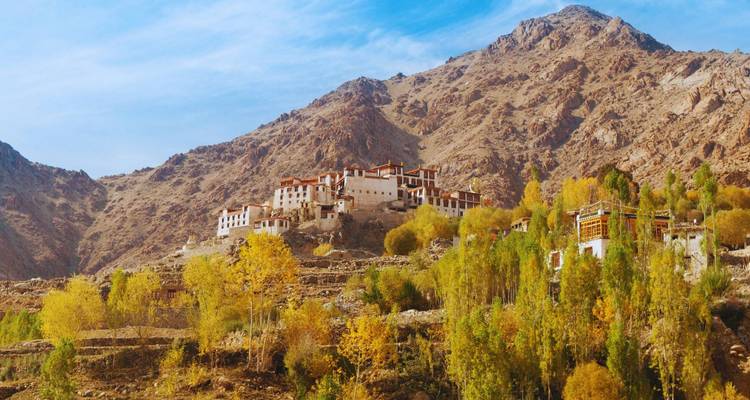 Un complejo monasterio en la ladera se eleva sobre árboles dorados de otoño contra montañas marrones escarpadas y un cielo azul.