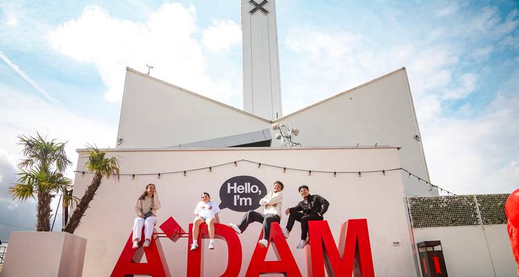 Junge Reisende sitzen auf riesigen roten Buchstaben, die "ADAM" buchstabieren, vor dem modernen A'DAM Tower in Amsterdam.