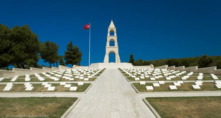 Cimetière militaire soigneusement aménagé avec drapeau turc et tour commémorative sous un ciel bleu profond ; filigrane présent.