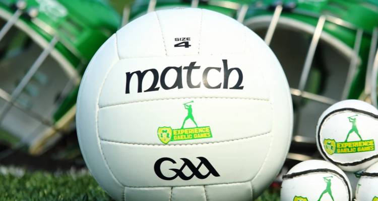 Primer plano de un balón de fútbol gaélico marcado con GAA y Experience Gaelic Games descansando sobre el césped junto a palos de hurling.