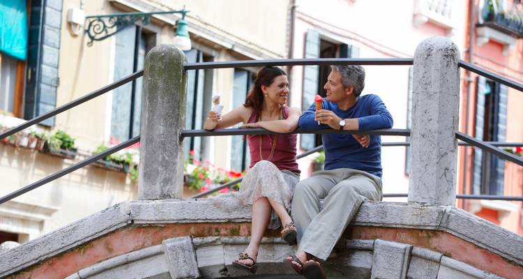 Couple sitting on a small Venetian bridge enjoying gelato and smiling at each other.