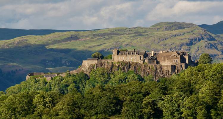 Historic hilltop castle surrounded by dense green forest and rolling Scottish hills lit by warm sunshine.