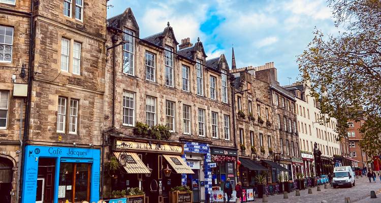 Historic stone storefronts and cafés lining a cobbled city street under a bright sky