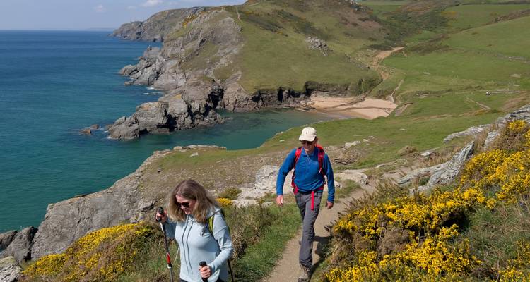Two hikers with trekking poles on a cliffside coastal trail overlooking turquoise sea and rugged cliffs