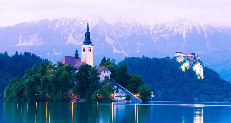 Iglesia de la isla del lago Bled y castillo reflejándose en aguas tranquilas con montañas nevadas al fondo