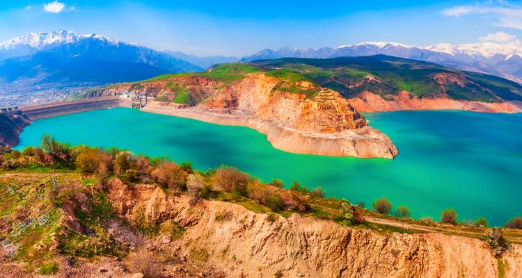 Réservoir turquoise brillant bordé de falaises rouges et de collines vertes avec des montagnes saupoudrées de neige au-delà.