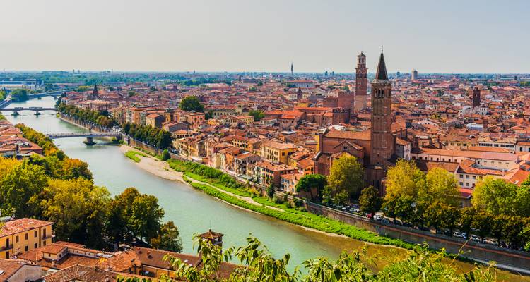 Wide river-side panorama of Verona with terracotta rooftops, medieval towers and lush riverside trees.
