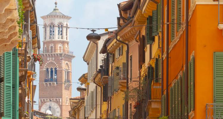 Colourful narrow street in Verona framed by shuttered houses with the Torre dei Lamberti visible in the distance.