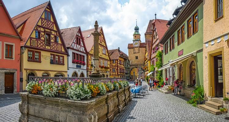 Bunte Fachwerkdorfstraße mit zentralem blumengefülltem Brunnen und Außensitzplätzen an einem hellen Tag.