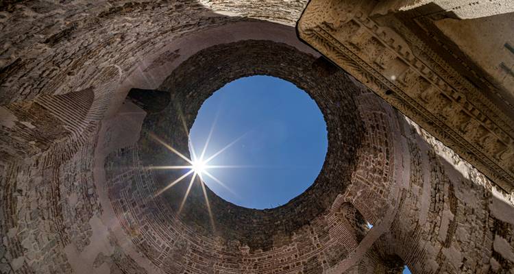 Vista hacia arriba a través de una abertura circular en antiguas paredes de piedra con el sol brillando como una estrella