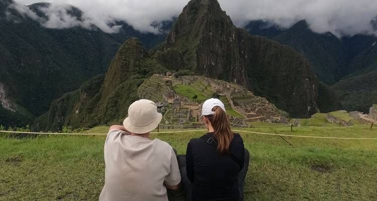 Deux voyageurs assis sur l'herbe contemplent l'ancienne citadelle de Machu Picchu entourée de montagnes brumeuses.