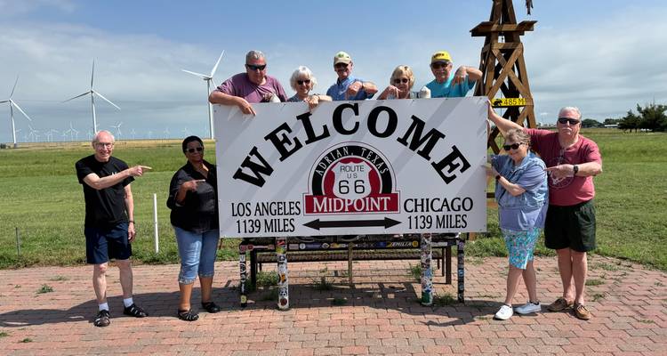 Grupo de turistas sonriendo alrededor del letrero del Punto Medio de la Ruta 66 que marca la distancia equidistante a Los Ángeles y Chicago.