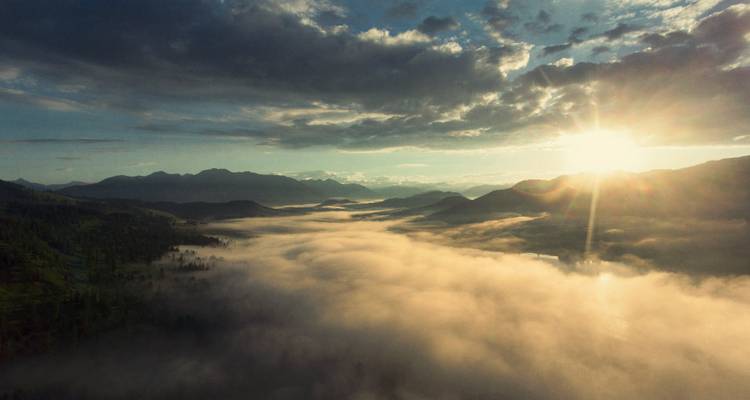 Luchtfoto van zonsopgang boven nevelvulde vallei met zonnestralen die door wolken priemen boven beboste bergen.