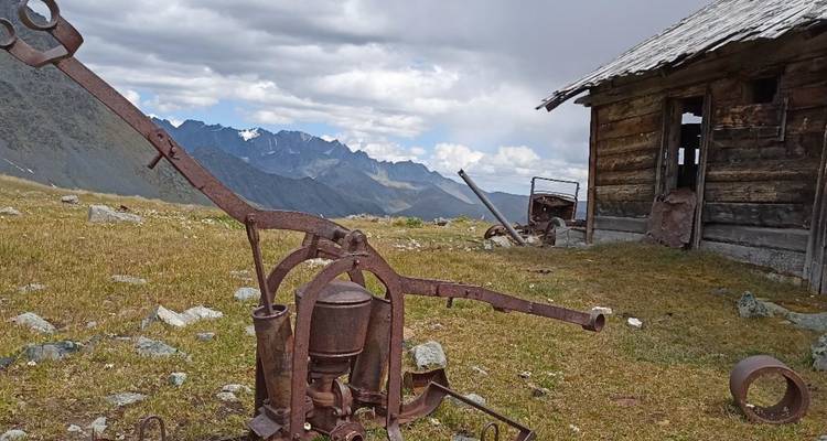 Machines abandonnées rustiques et cabane en bois situées sur un pré alpin avec des pics déchiquetés au loin.