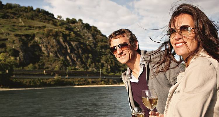 Glimlachend stel met wijnglazen genietend van een schilderachtige riviercruise langs groene heuvels en rotsachtige wijngaarden.