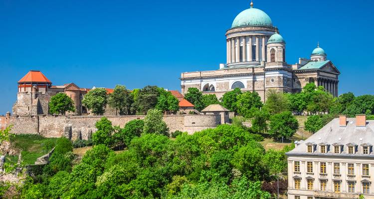 De Basiliek van Esztergom torent boven groene boomkruinen en historische fortmuren uit tegen een diepblauwe lucht.
