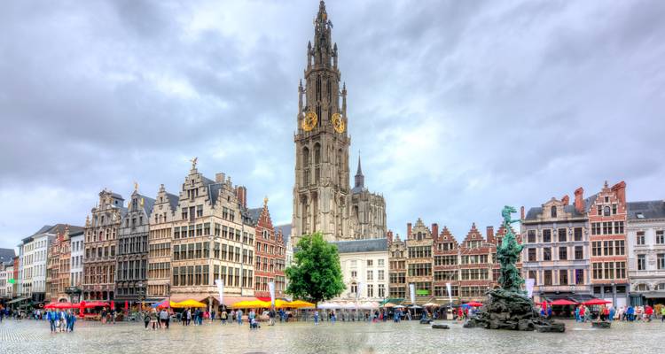 Historische marktplein in Antwerpen met sierlijke gildenhuizen, katedraalspits en kleurrijke parasols rond de fontein op een bewolkte dag.