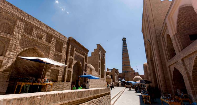 Ruelle ensoleillée de Khiva avec une architecture de brique et un minaret rayé imposant au loin.
