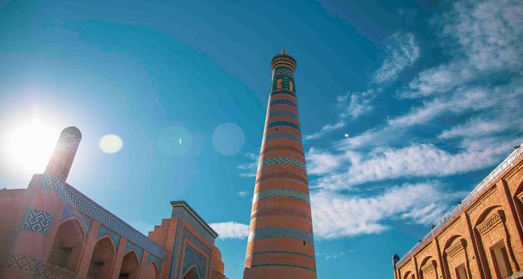 Vue en contre-jour du minaret emblématique de Khiva s'élançant dans un ciel bleu profond avec des reflets de soleil.