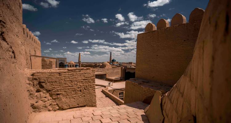 Vue ensoleillée le long de remparts en briques de terre crue donnant sur une vieille ville avec un dôme turquoise lointain et un minaret sous un ciel bleu profond et des nuages épars.