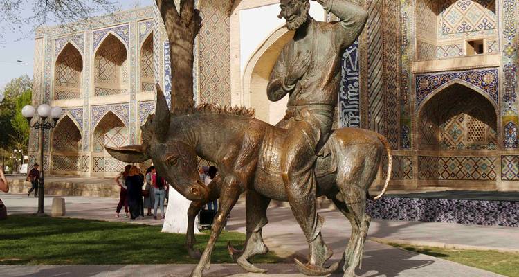 Statue de bronze d'un homme barbu chevauchant un âne placée devant la façade richement carrelée d'une médersa, avec de petits groupes de visiteurs à proximité.