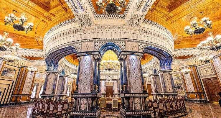Weelderige paleiszaal met prachtige kroonluchters, een uitgebeeldhd houten plafond en rijkelijk versierde zuilen die een ingewikkelde ronde galerij vormen.