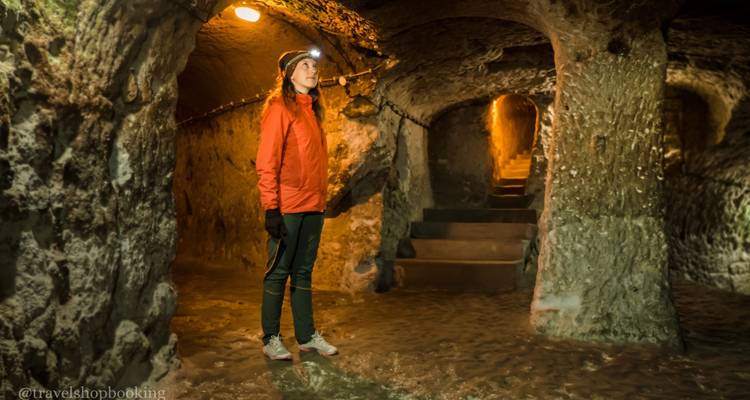 Una mujer con una chaqueta roja explora una ciudad subterránea de cuevas iluminada, parada sobre piedra húmeda junto a escaleras talladas.