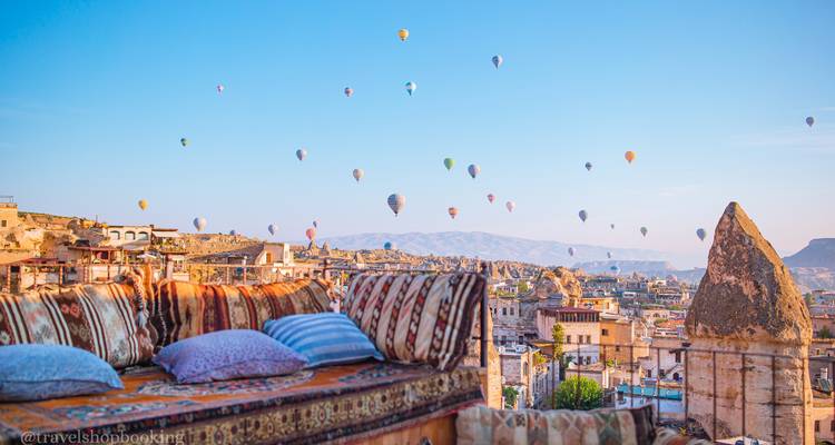 Cojines coloridos en una terraza en la azotea contemplan el paisaje urbano de Capadocia mientras docenas de globos aerostáticos se elevan en el cielo matutino despejado.