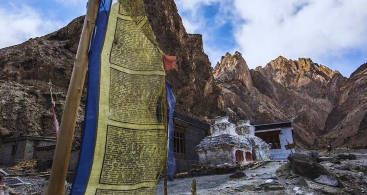 Eine wettergezeichnete buddhistische Gebetsfahne flattert vor schroffen braunen Bergen und kleinen weißen Stupas in einem abgelegenen Himalaya-Tal.