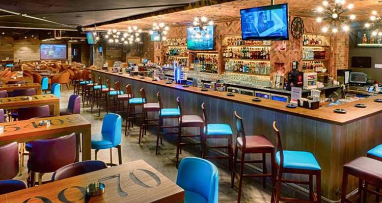 Bar de hotel elegante con mostrador largo de madera, sillas coloridas y múltiples pantallas de televisión.