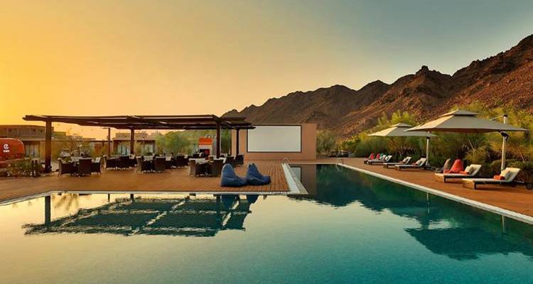 Piscina infinita junto a montañas al atardecer dorado con área de comedor al aire libre y tumbonas.