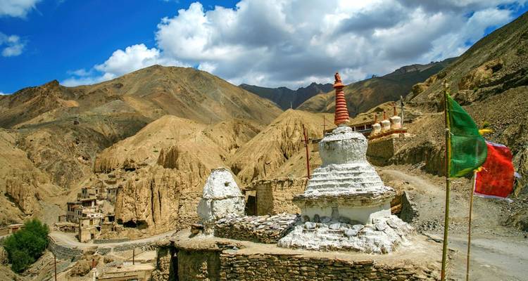 Weiße Chörtens und Gebetsfahnen vor kargen ockerfarbenen Hügeln und verstreuten Steinbehausungen unter einem strahlend blauen Himmel.