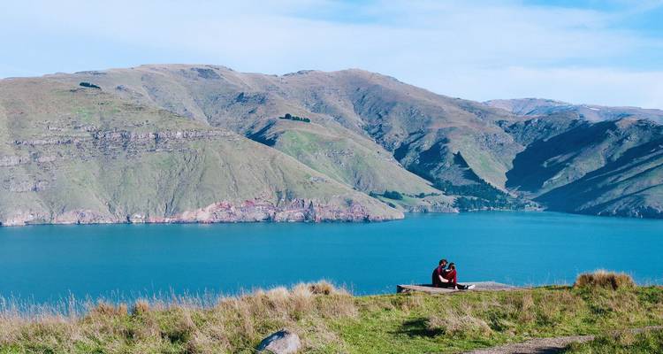 Ein Paar sitzt auf einer Bank mit Blick auf einen weiten blauen See, umgeben von sanften grünen Hügeln unter einem klaren Himmel.