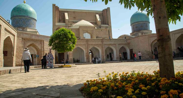 Cour intérieure du complexe Khast-Imam avec des dômes bleus jumeaux, des parterres fleuris et des visiteurs se promenant au soleil.