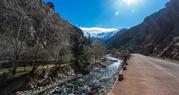 Kristalheldere beek stroomt naast een bergweg die leidt naar besneeuwde toppen van de Hoge Atlas onder een heldere zon.