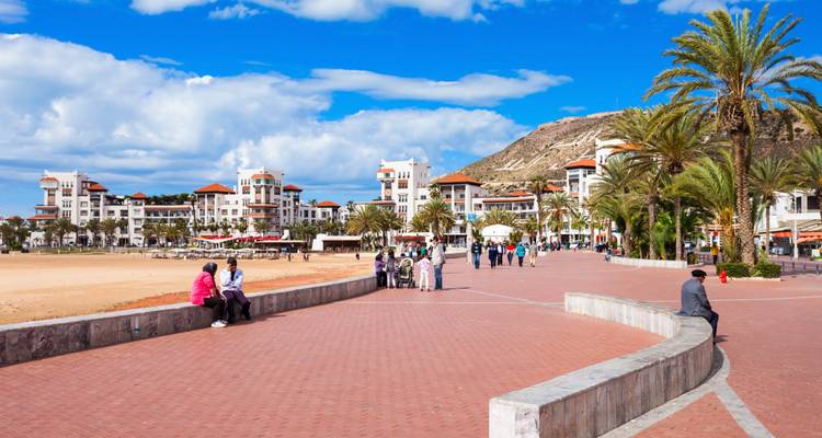 Palmboomgezoomde zeeboulevard in Agadir met vakantiegangers die wandelen langs het zandstrand en resorthotels.