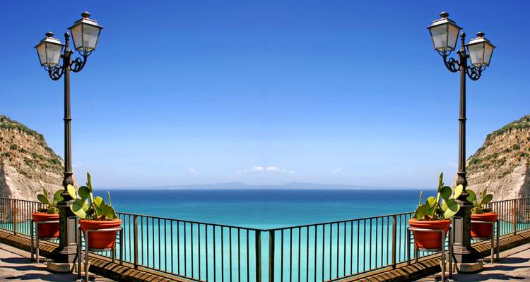 Symmetrisch ijzeren balustrade uitkijkpunt met cactussen in potten met uitzicht op kalme azuurblauwe zee en verre horizon.